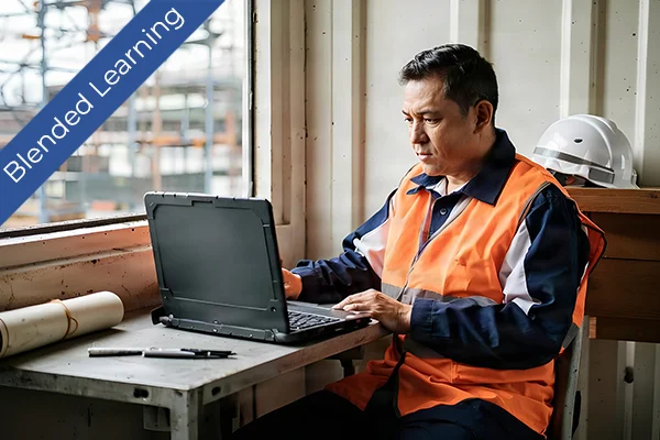 A worker in an orange safety vest using a laptop at a construction site office.