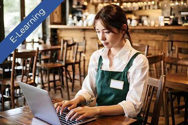 A woman in an apron uses a laptop for online health and safety training in a cafe.