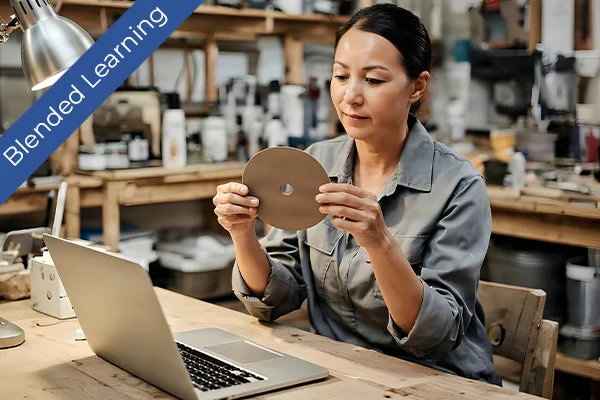 A professional in a workshop examines an abrasive disc beside a laptop, illustrating practical application in blended learning.